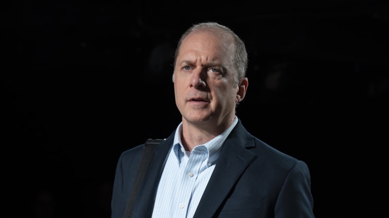 An older white man wears a navy blazer over a light blue striped collared shirt tucked into khakis. A black messenger bag hangs on his shoulder.